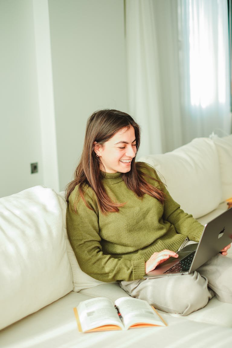 A young woman enjoys working on her laptop and blog while relaxing on a sofa indoors, surrounded by books.