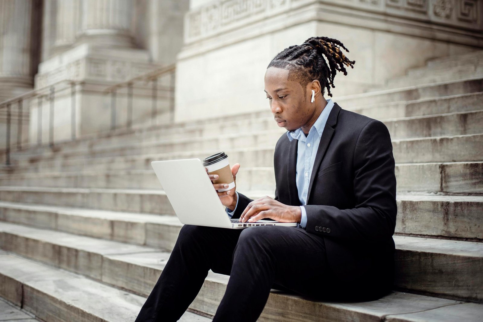 Concentrated African American businessman wearing formal clothes with coffee to go and earbuds browsing netbook while blogging remotely on stone building stairs