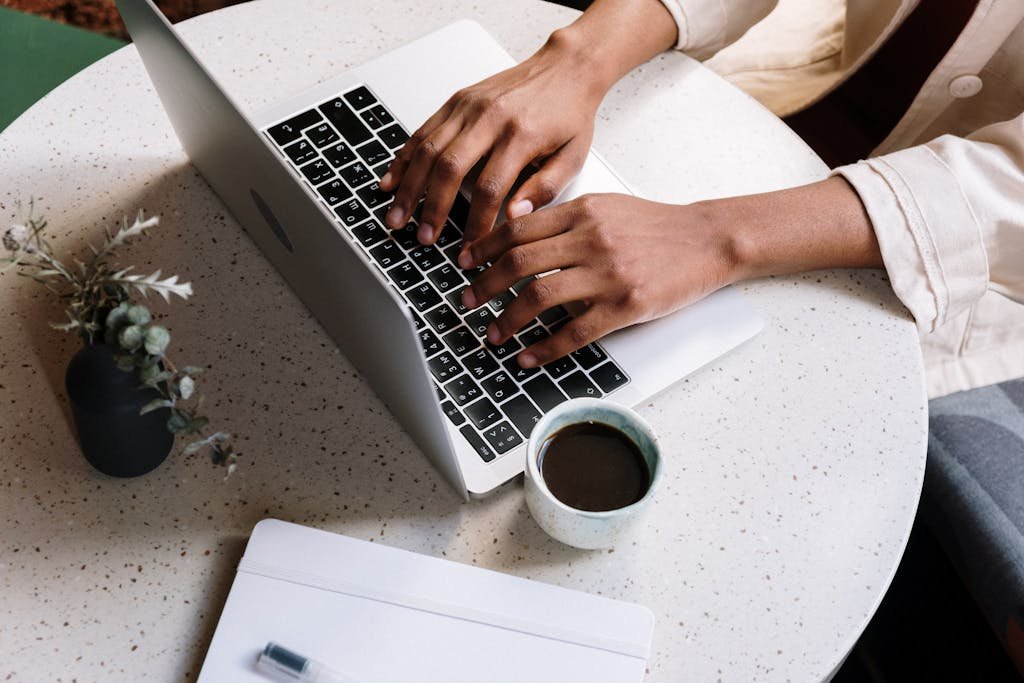 A person typing on a laptop at a café table with coffee and a notebook working blog
