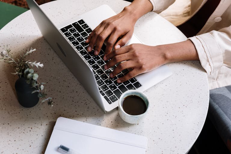 A person typing on a laptop at a café table with coffee and a notebook working blog