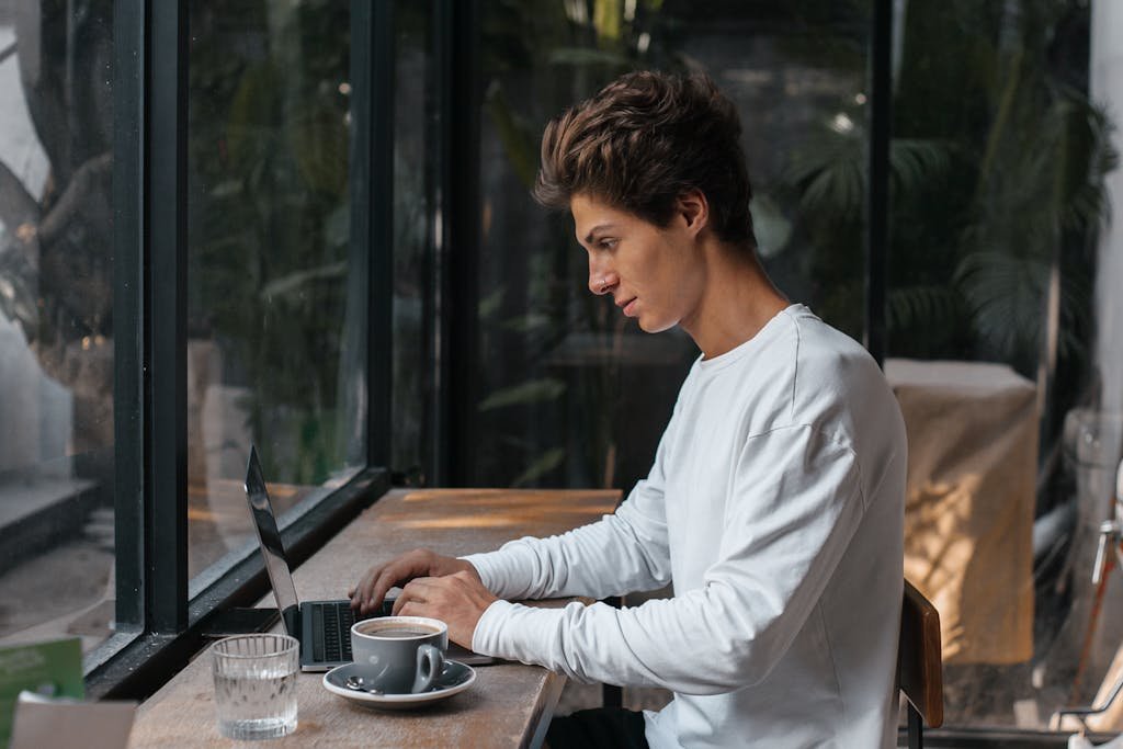 A young man in a white sweater working on a laptop in a cozy cafe with large windows.
