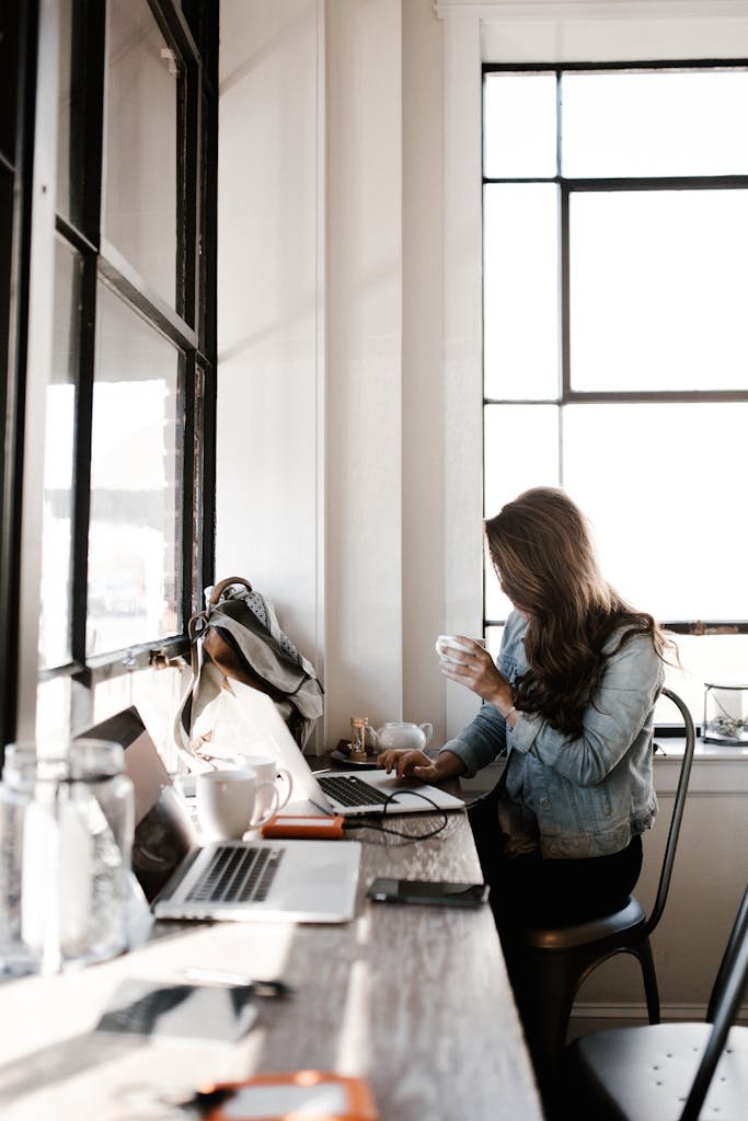 A young woman working on her laptop, sipping coffee in a bright café setting, brainstorming blog post ideas.