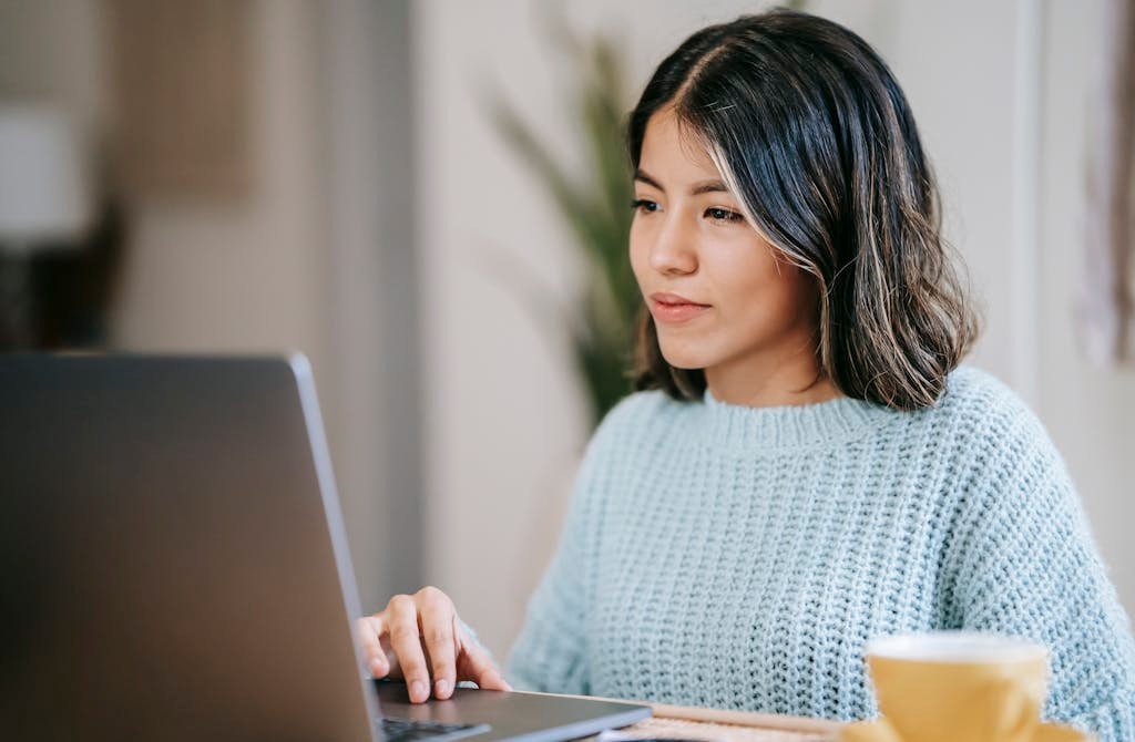 Confident young Latin American woman in casual clothes working remotely on netbook at table near mug with coffee working on how to start a blog and make money