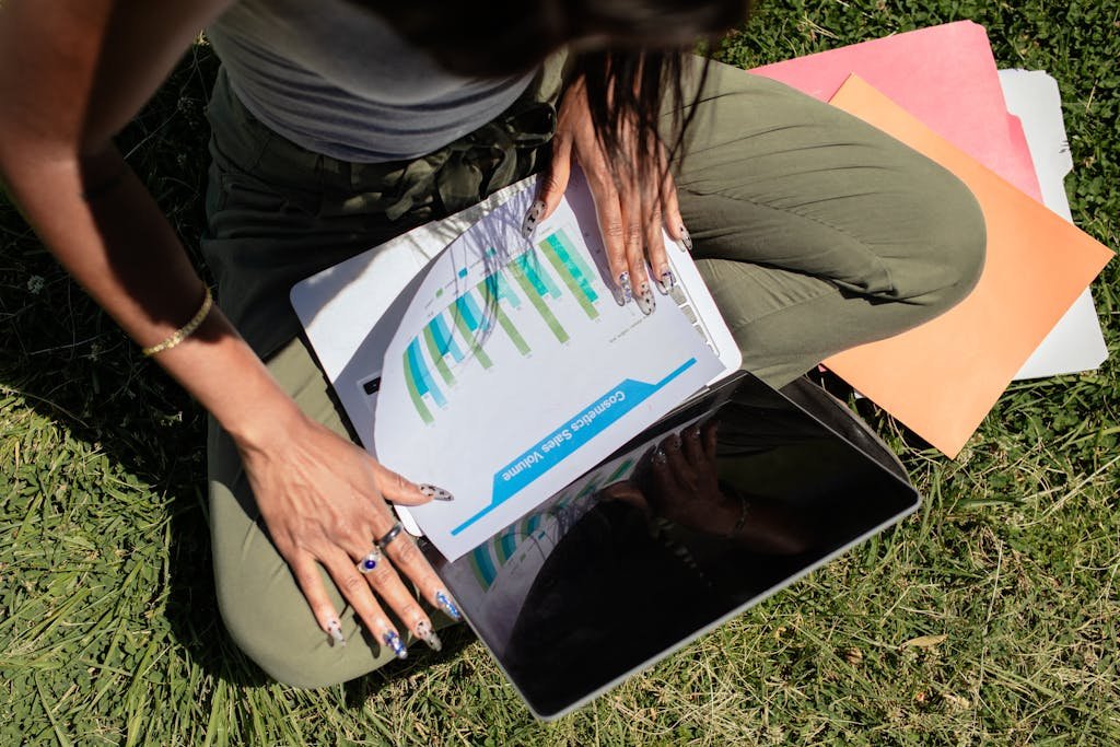 Woman working outdoors on grass with a laptop and documents, reviewing her digital product funnel