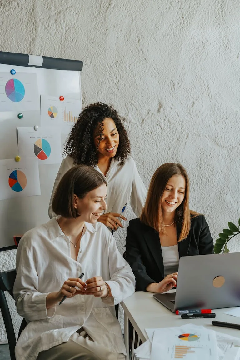 Three businesswomen collaborate with graphs and a laptop in a modern office setting working on best AI prompts